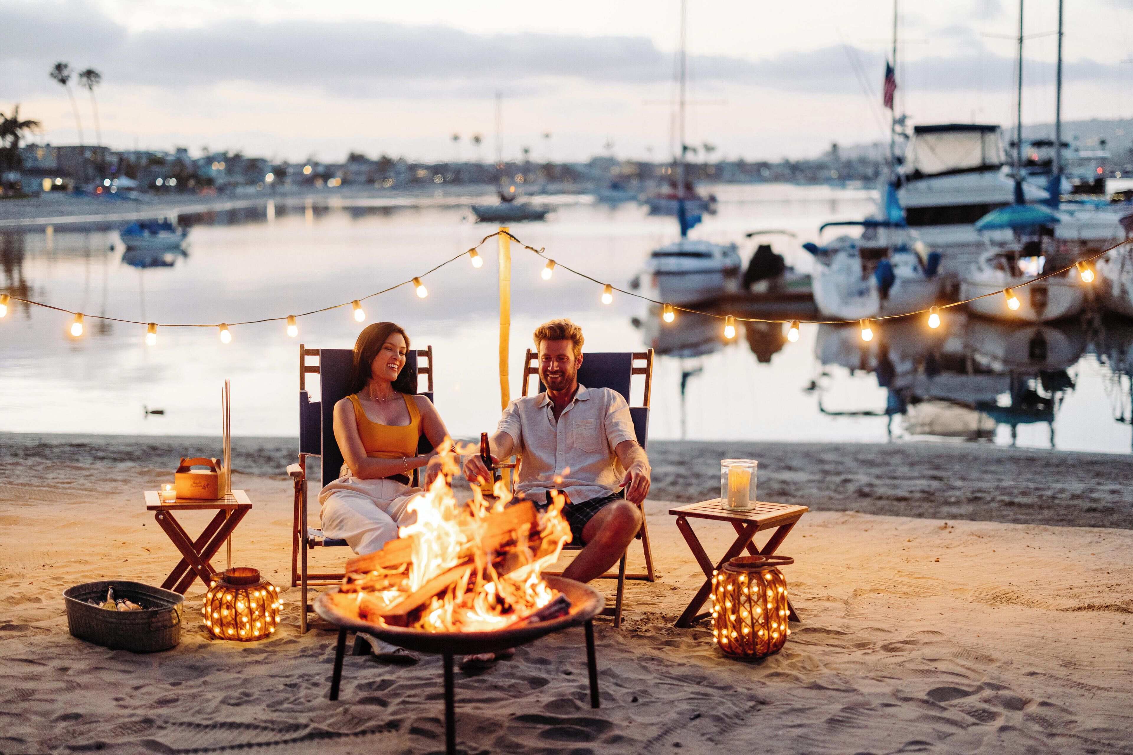 Ein Paar entspannt am Strand bei einem Lagerfeuer mit Lichterketten und blickt auf einen Hafen mit Booten.