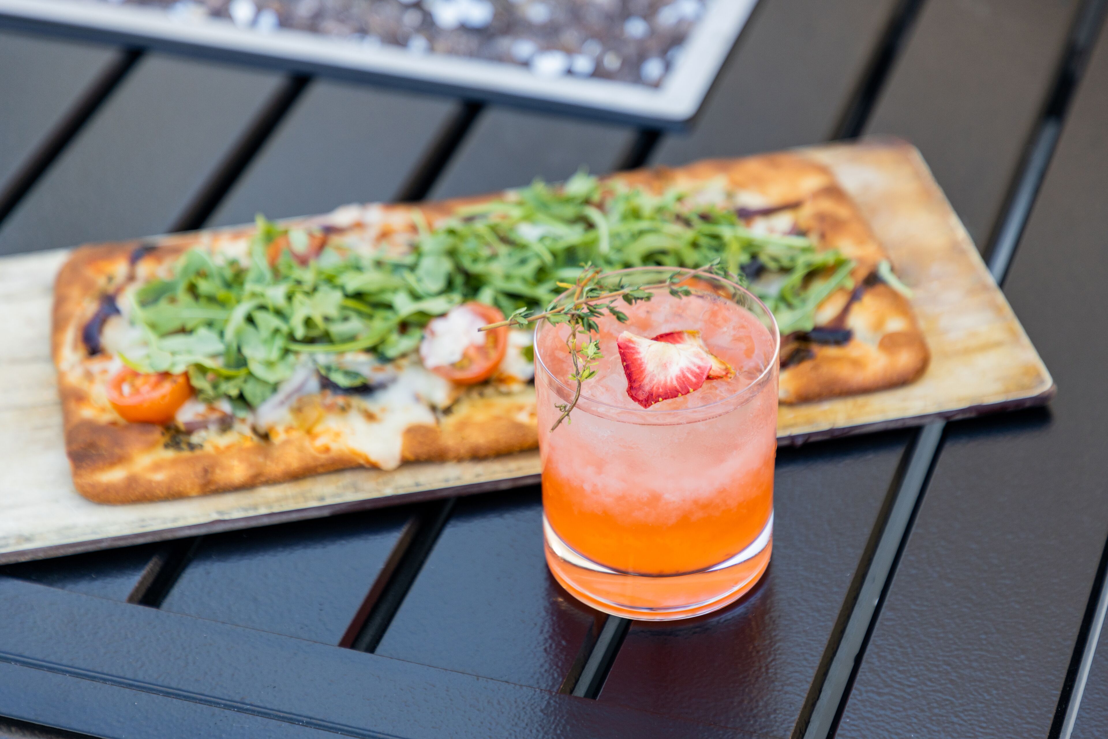 A rectangular pizza with greens and tomatoes on a wooden board, and a glass with a red drink and a strawberry on a black table.