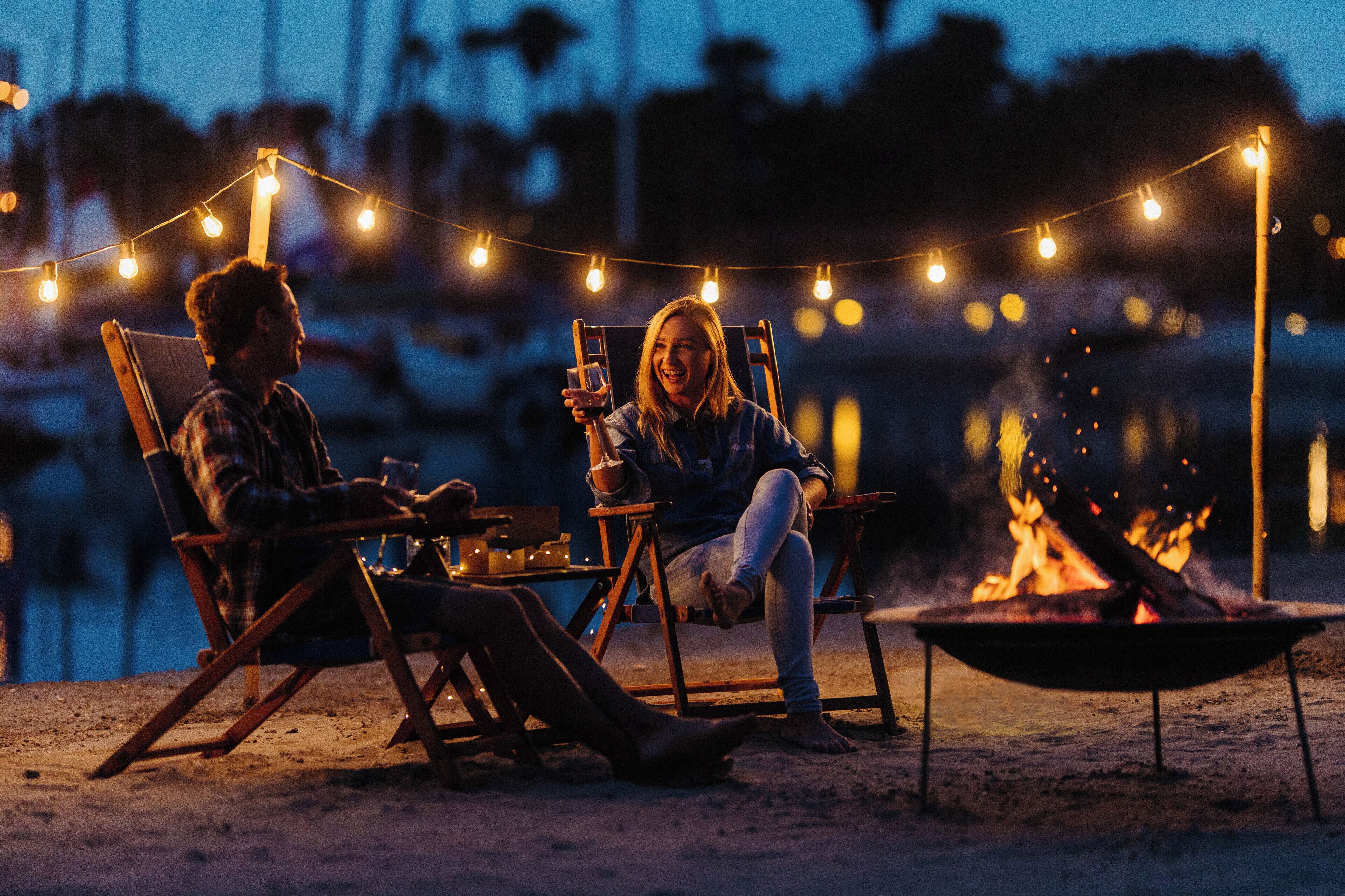 Two people sitting on wooden chairs outdoors at night, with string lights overhead and a campfire in the foreground.