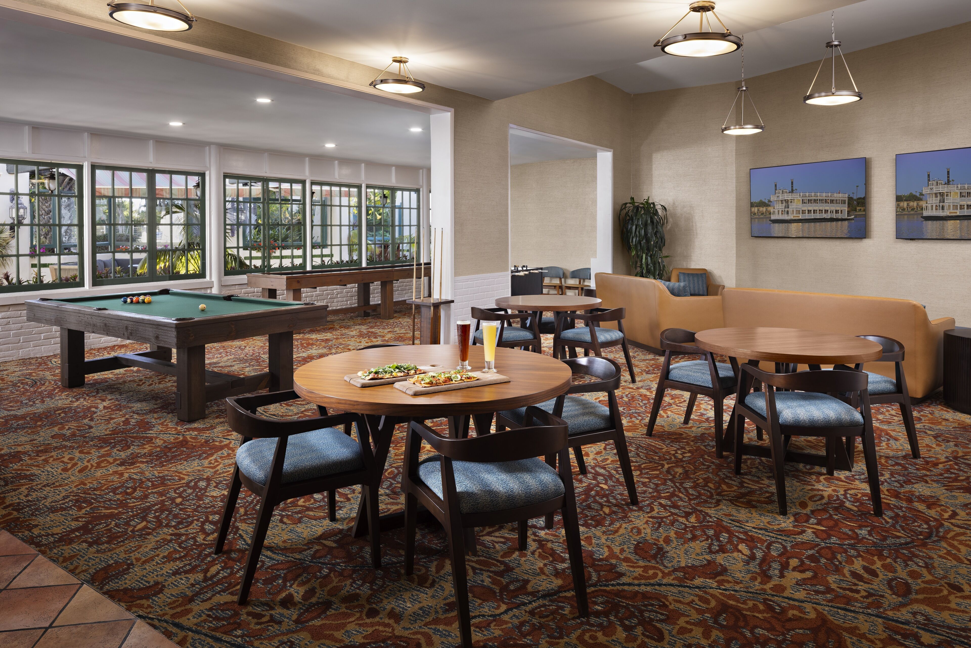 A pool table, billiards balls, and a pool cue in a room with a patterned carpet, a round table with chairs, and a sofa.
