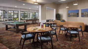 A pool table, billiards balls, and a pool cue in a room with a patterned carpet, a round table with chairs, and a sofa.