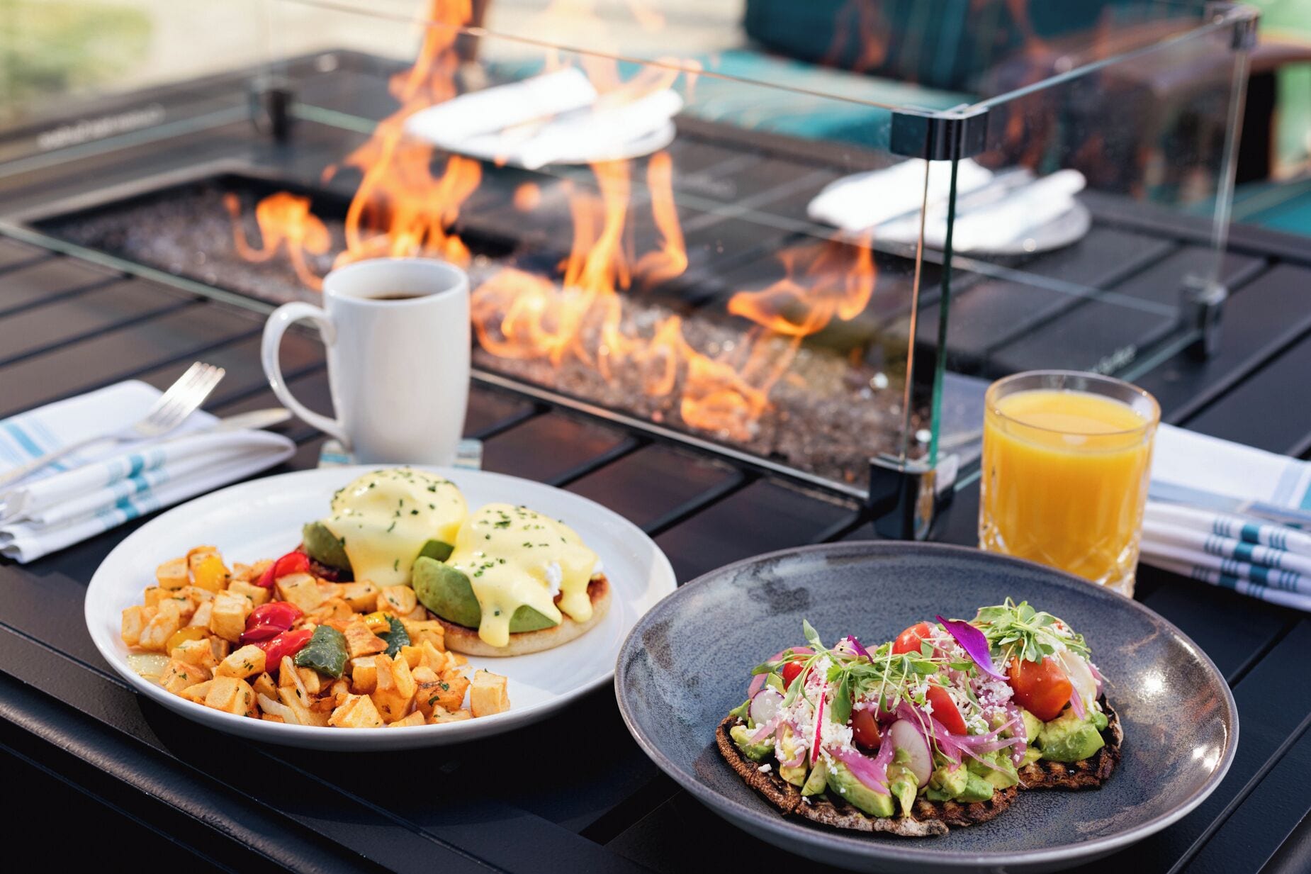 A meal setup with a plate of eggs benedict, a bowl of roasted vegetables, and a toast with avocado and vegetables, with a glass of orange juice and a cup of coffee on a table with a fire feature in the background.