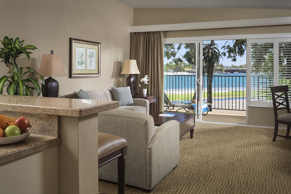 Une chambre d’hôtel avec un coin salon, une table à manger avec des fruits, une plante et une vue sur la plage.
