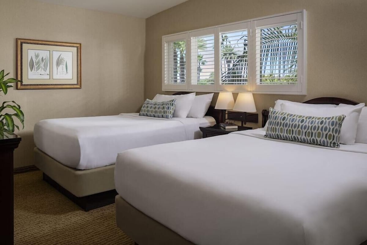 Quarto de hotel com duas camas, uma mesa de cabeceira com abajur, uma impressão botânica emoldurada na parede e uma janela com vista para vegetação.