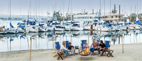 A marina with boats docked, a fire pit with people around it, and a building with a tower in the background.