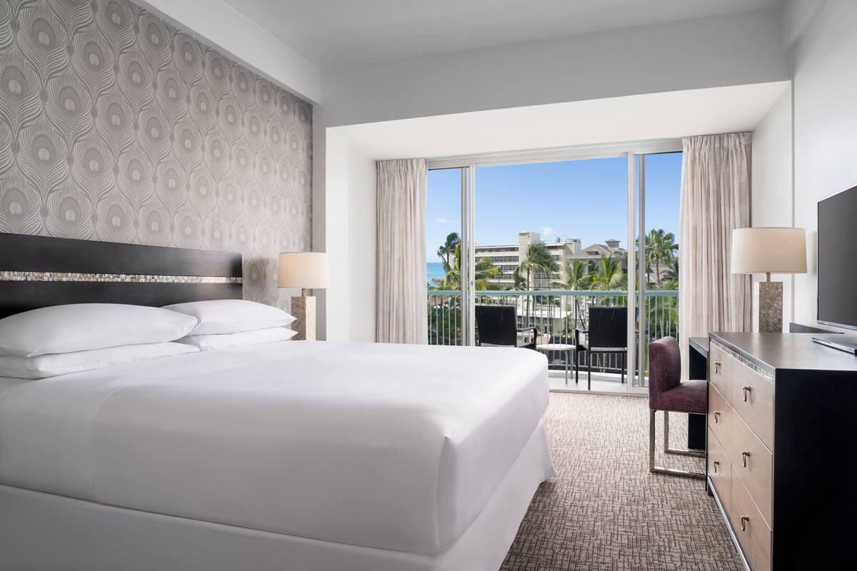 A hotel room with a large bed, a desk, a chair, a TV, and a balcony with a view of palm trees and buildings.