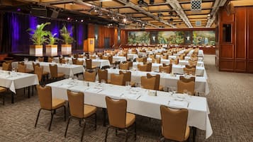 A banquet hall with multiple tables set for dining, a stage with plants, and a mural on the wall.