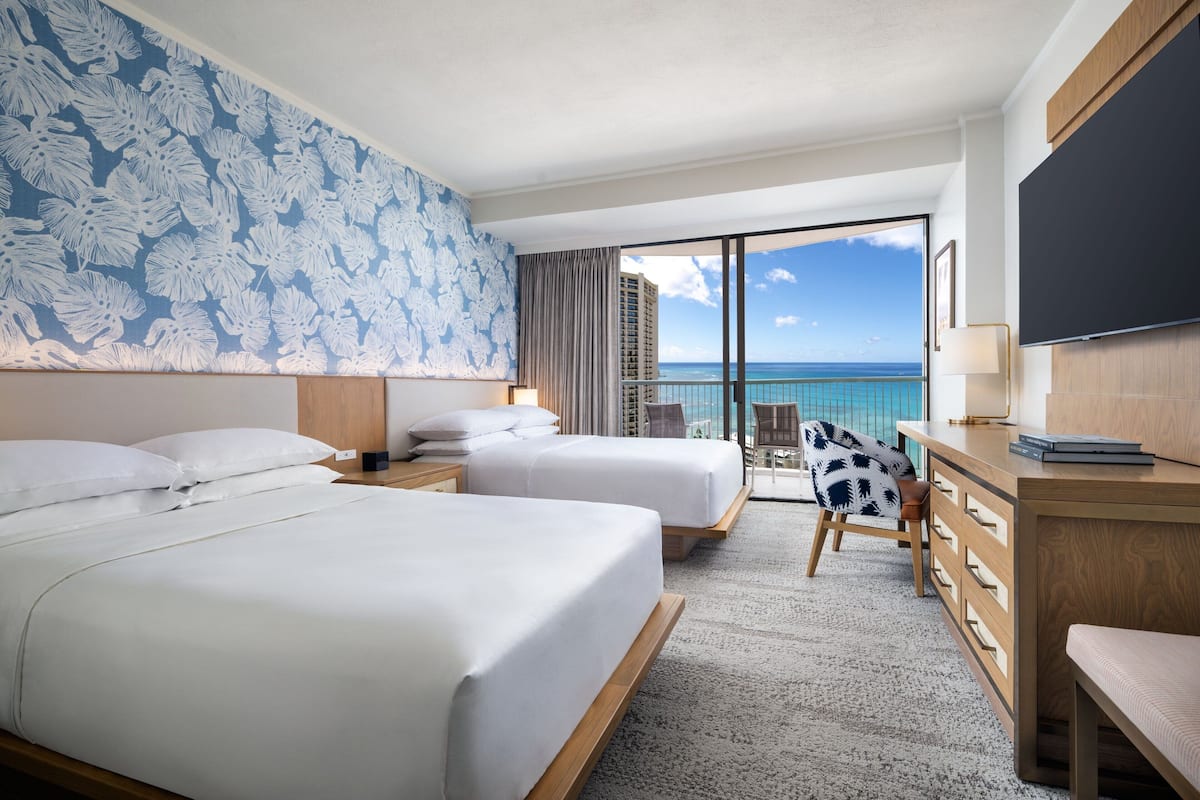A hotel room with two beds, a desk, a chair, and a large window with a view of the ocean.