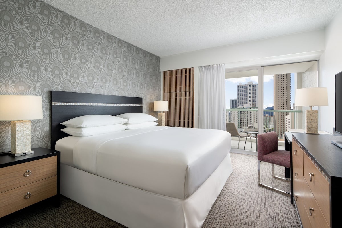 Habitación de hotel con una cama grande, dos mesitas de noche con lámparas, un escritorio con una silla y vistas a la ciudad a través de amplios ventanales.