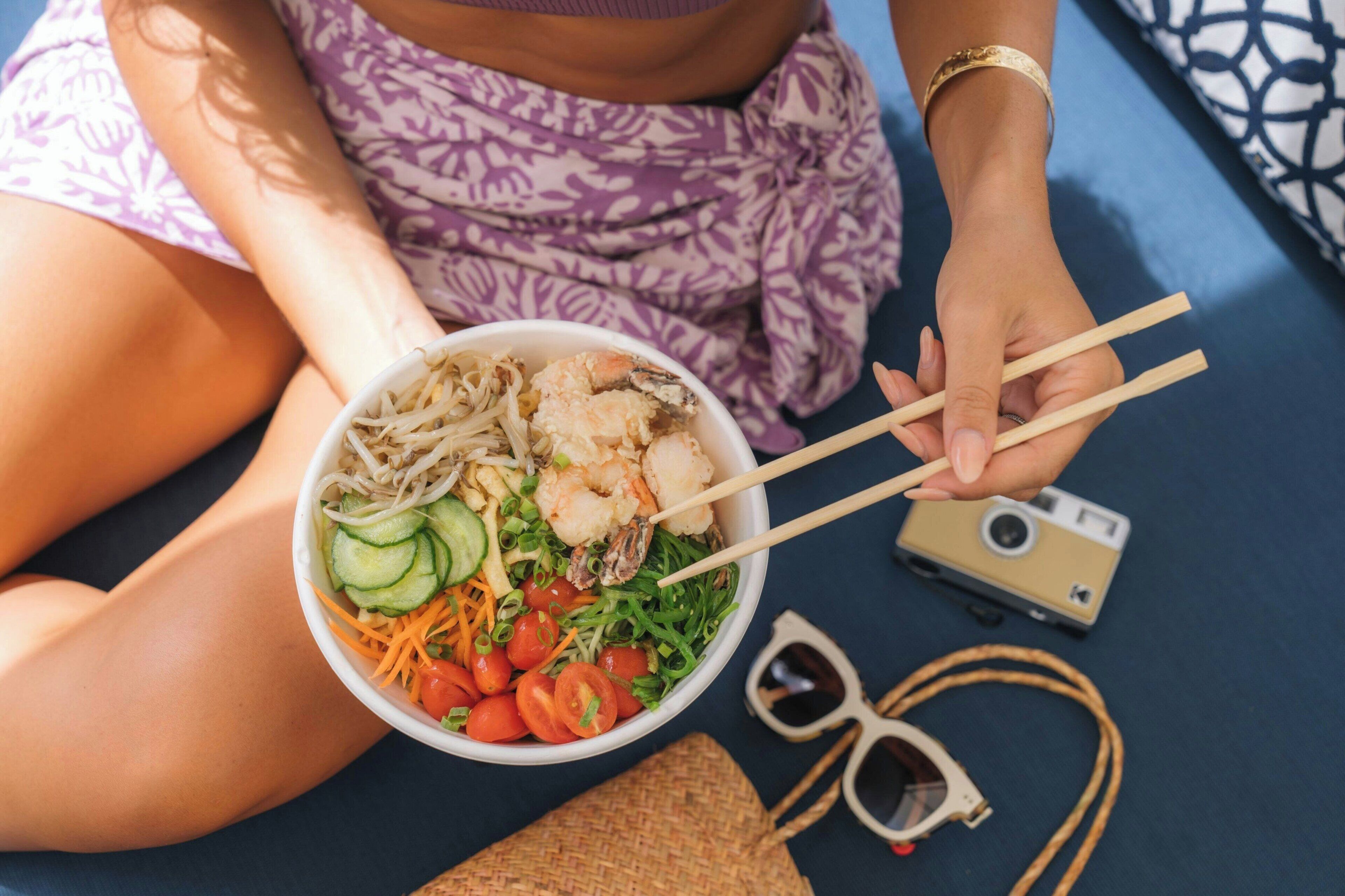 A person holding a bowl of salad with chopsticks, a camera, and sunglasses on a table.
