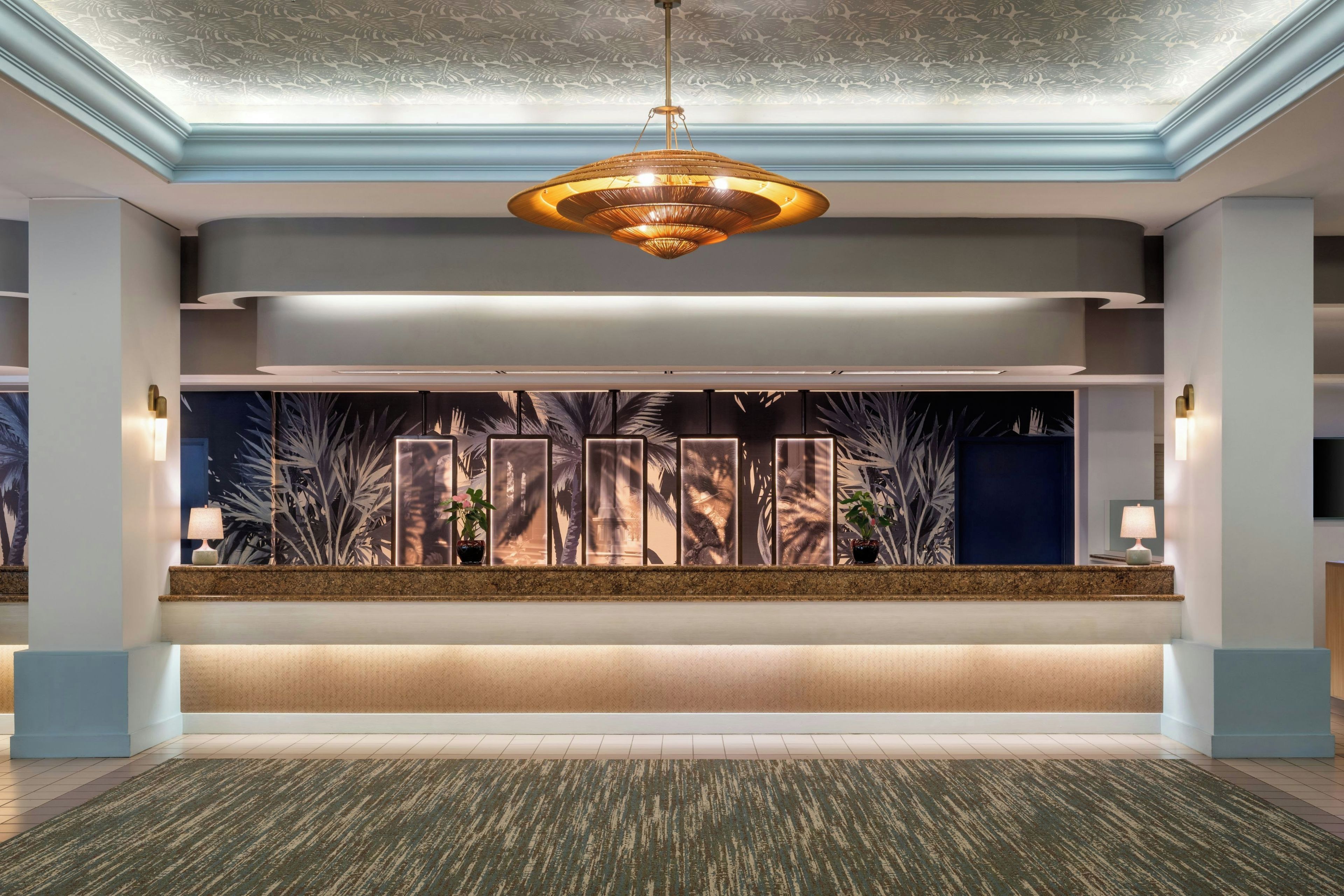 A hotel lobby with a central reception desk, a large hanging light fixture, and a patterned wall with plants.