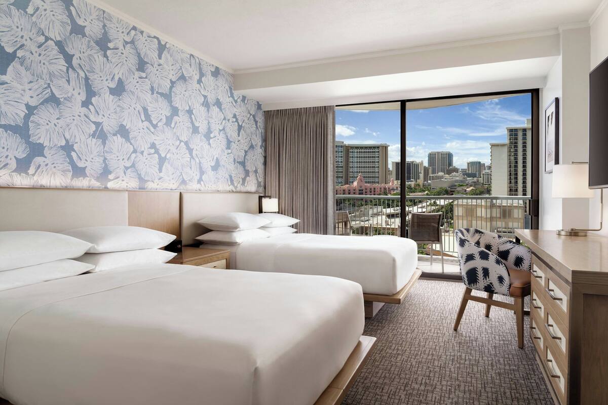 A hotel room with two beds, a desk, and a view of the cityscape.