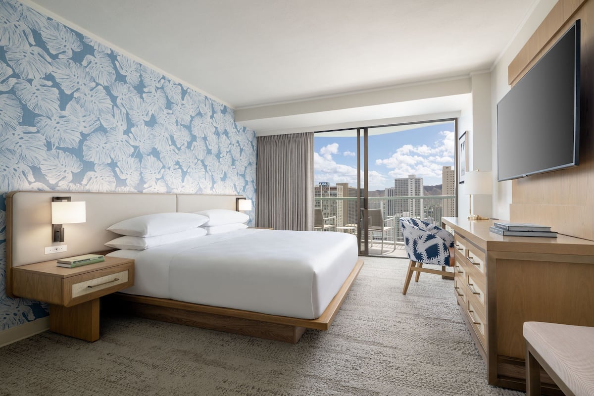 A hotel room with a large bed, a desk, a chair, a TV, and a balcony with a city view.