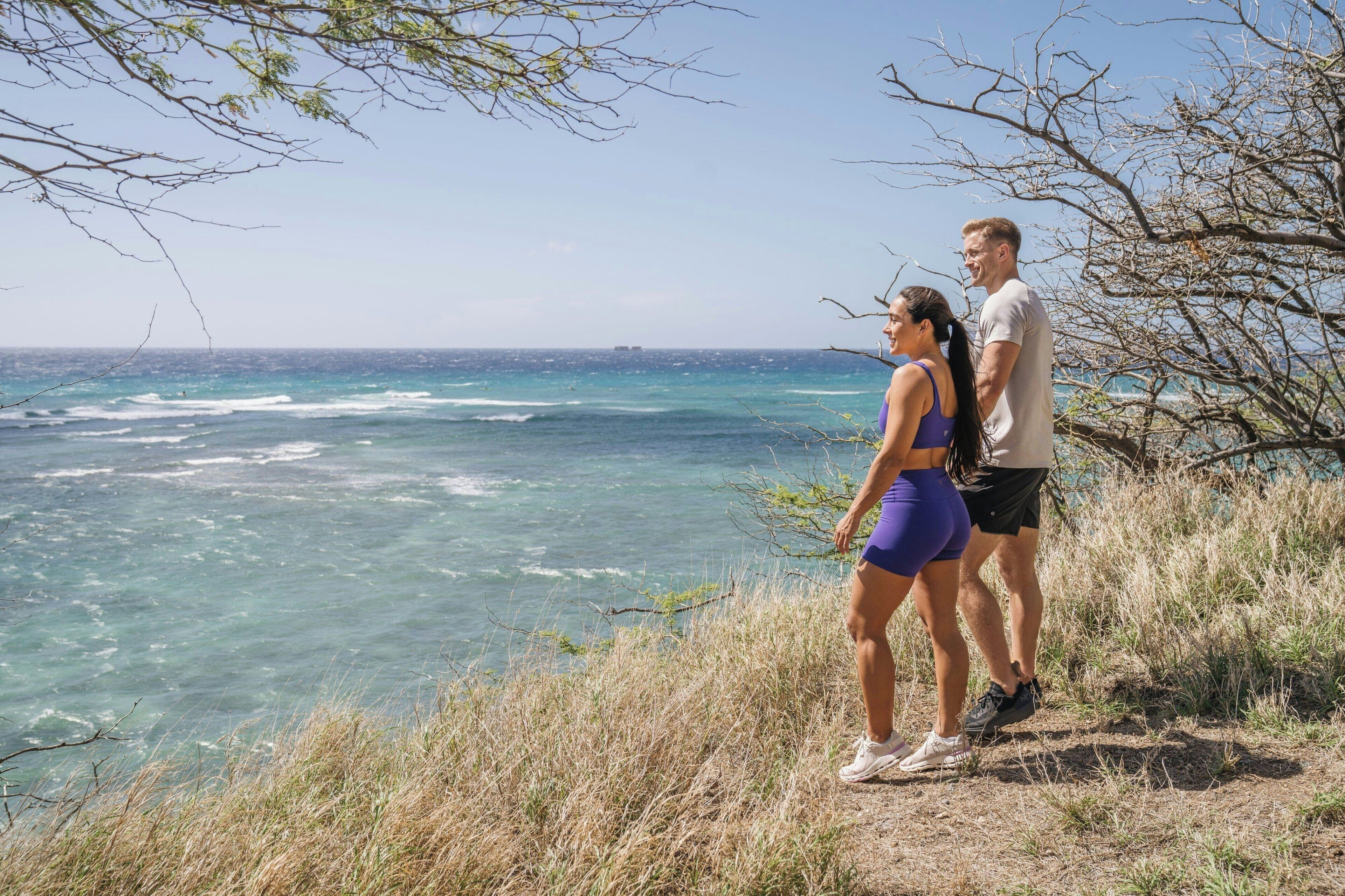 A couple standing on a grassy cliff overlooking a coastal view with the ocean and a distant ship.