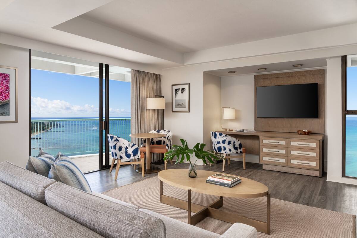 A modern hotel room with a balcony, a sofa, a coffee table, a TV, and a desk with a lamp.