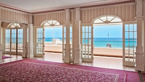 Una habitación amplia con un gran tapete rojo estampado, columnas blancas y varias puertas que dan a un balcón con vistas al océano.