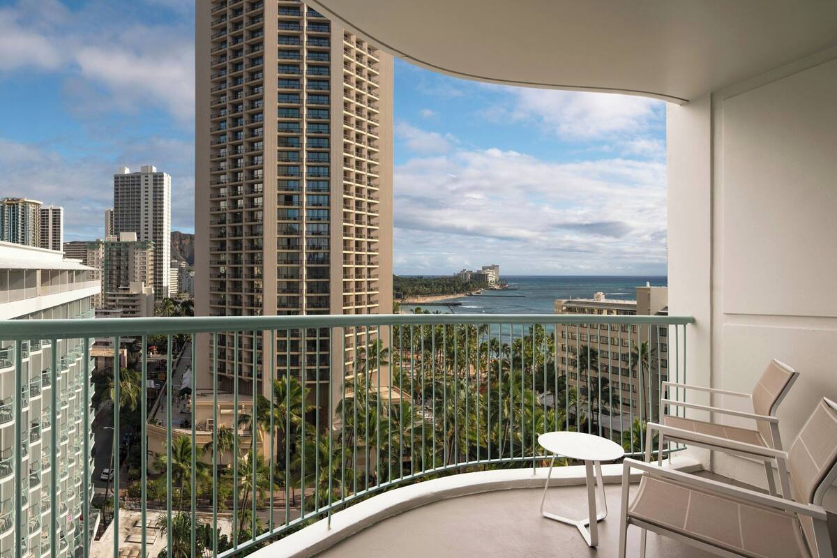 A balcony with a curved railing, a small table, and two chairs. A tall building with a grid-like facade is visible in the background.