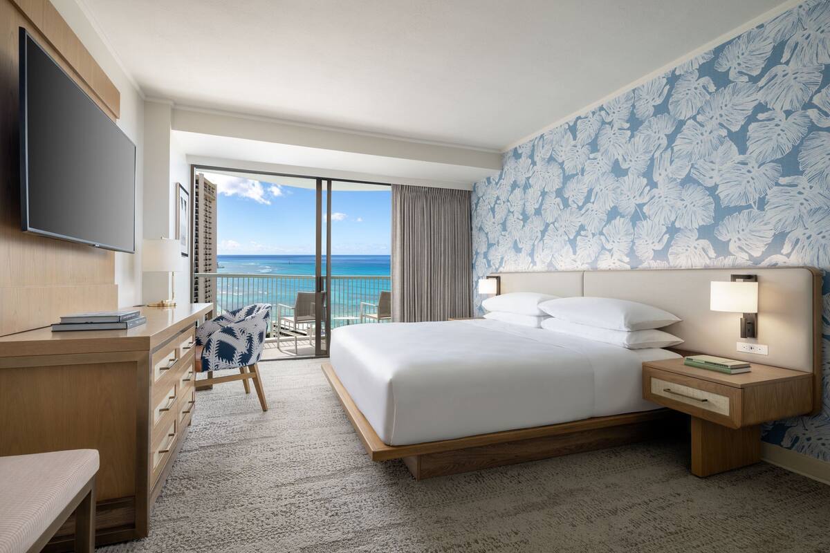 A hotel room with a large bed, a desk, a chair, a TV, and a balcony with a view of the ocean.
