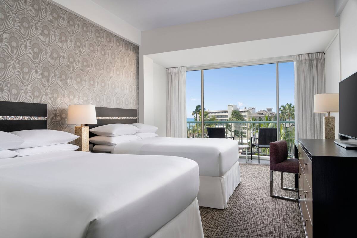 A hotel room with two beds, a desk, a chair, and a large window offering a view of buildings and palm trees.