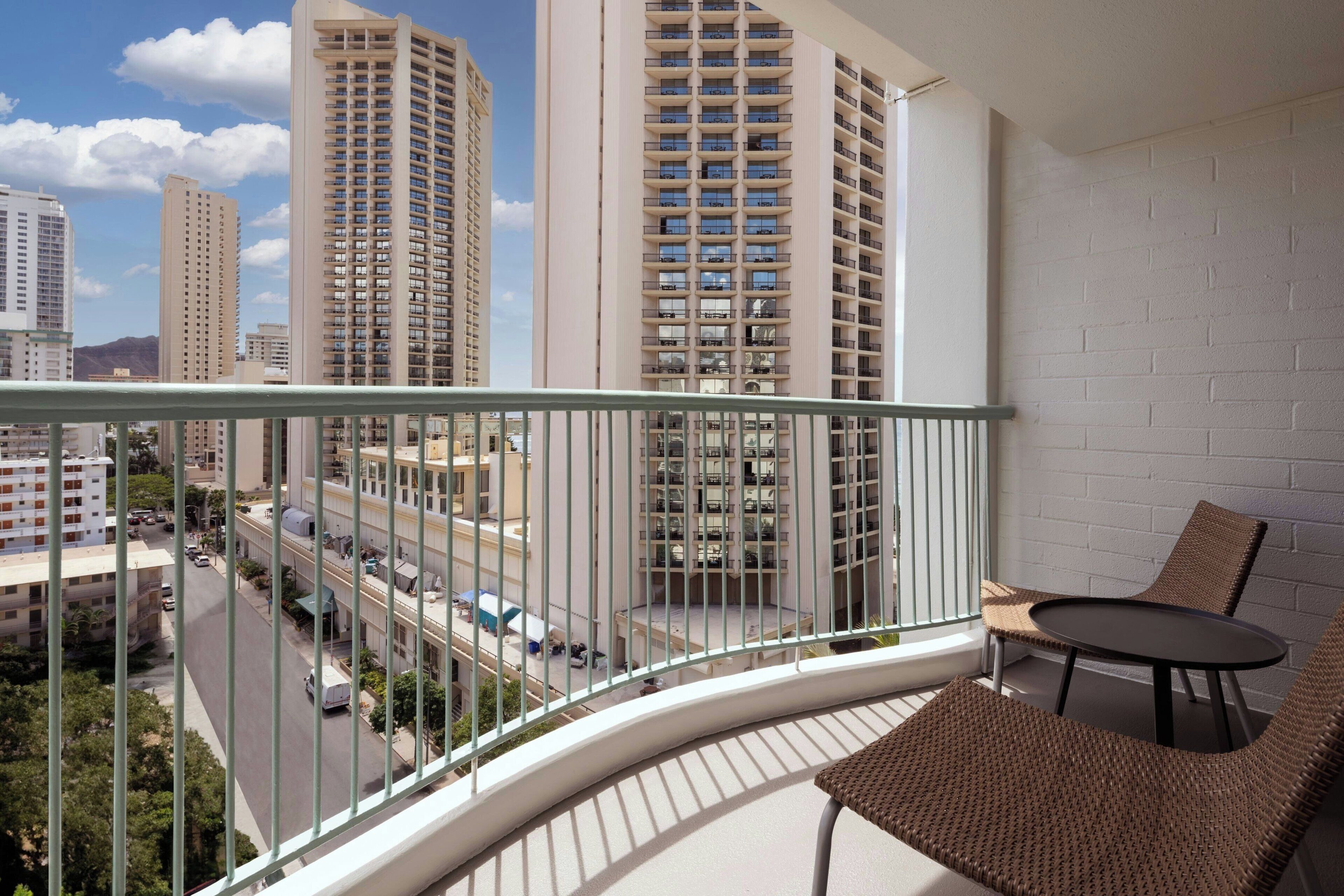 Un balcon avec une chaise et une table en osier, donnant sur une vue panoramique de la ville, avec ses nombreux immeubles.