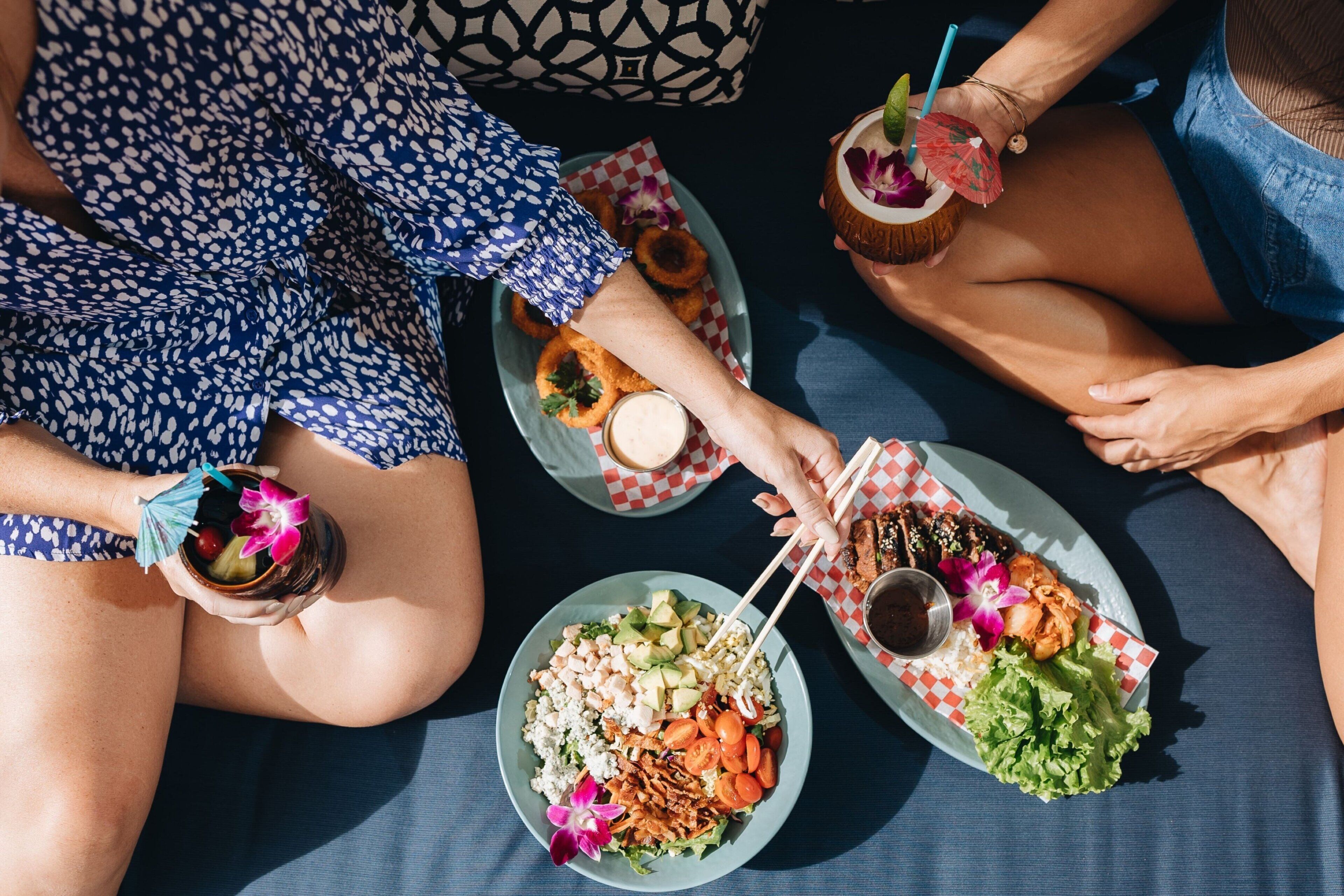 A group of people sitting on a blue mat with plates of food, drinks, and a coconut drink with a straw.