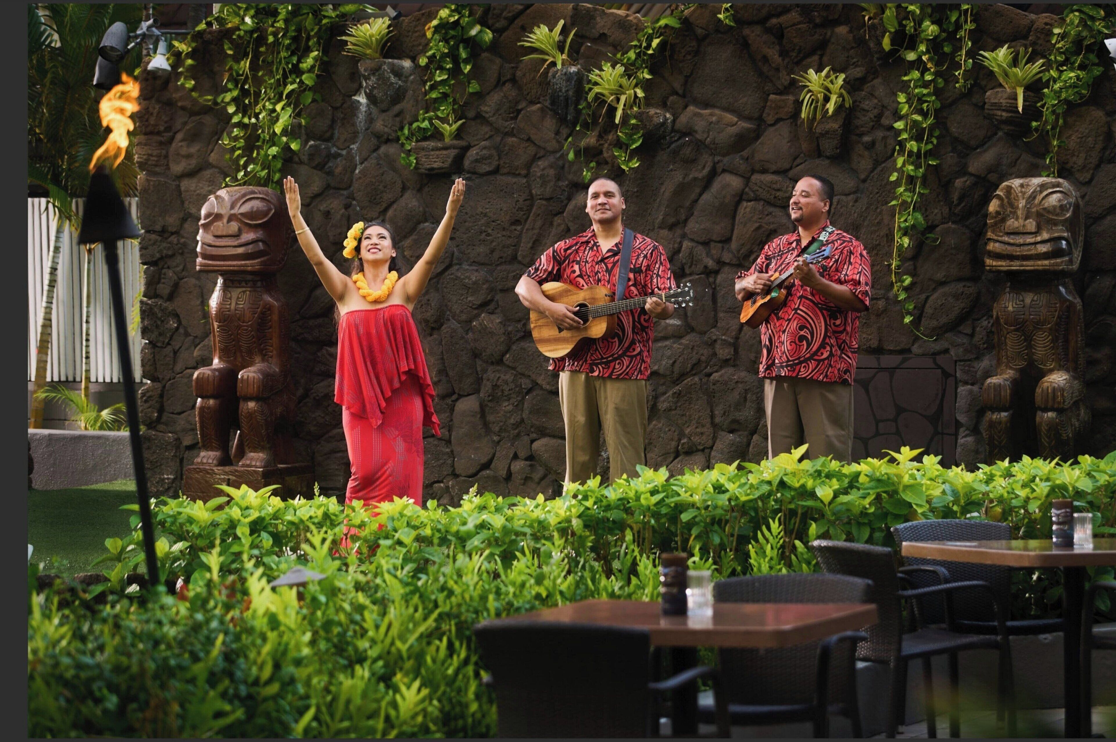 Un escenario con tres artistas, un antorcha tiki y estatuas de tiki de piedra en un entorno exterior.