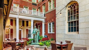 An outdoor dining area with tables and chairs, a statue, and a brick building with windows. - Sheraton Commander Hotel (Cambridge)