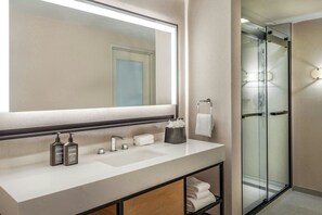 A modern bathroom with a large mirror, a white sink, two bottles of shampoo, a glass shower enclosure, and a towel rack.
