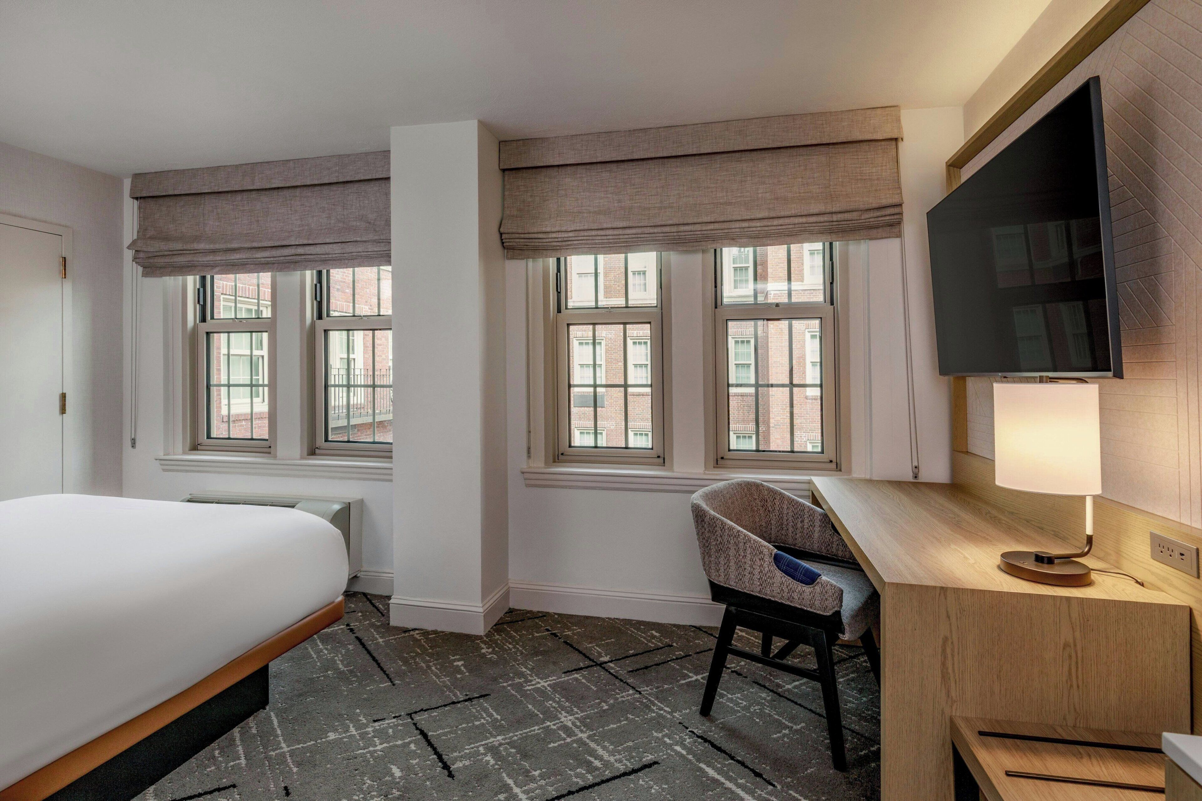A hotel room with a bed, a desk with a chair, a TV, and large windows.