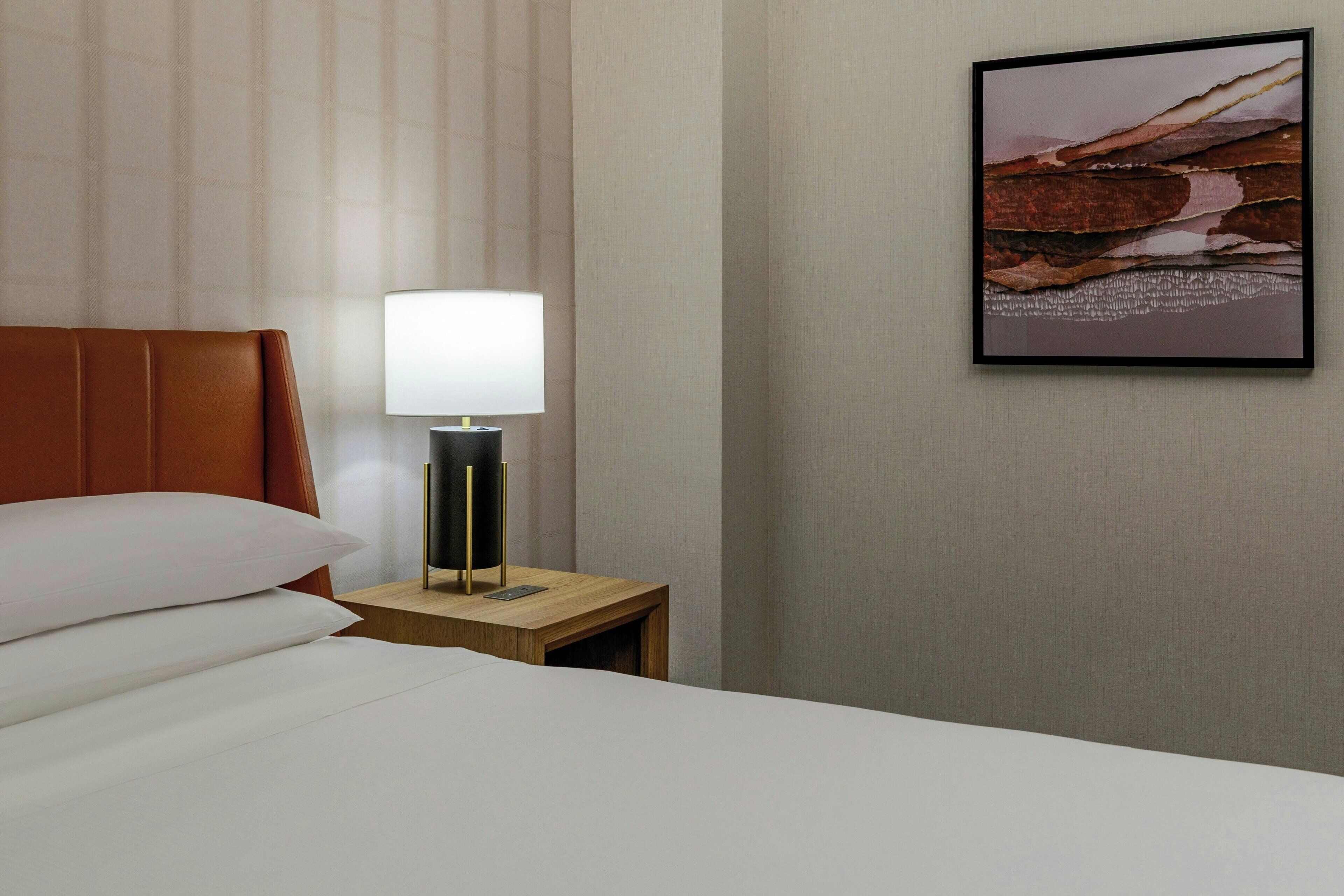 A hotel room with a bed, pillows, a nightstand with a lamp, and a framed picture on the wall.