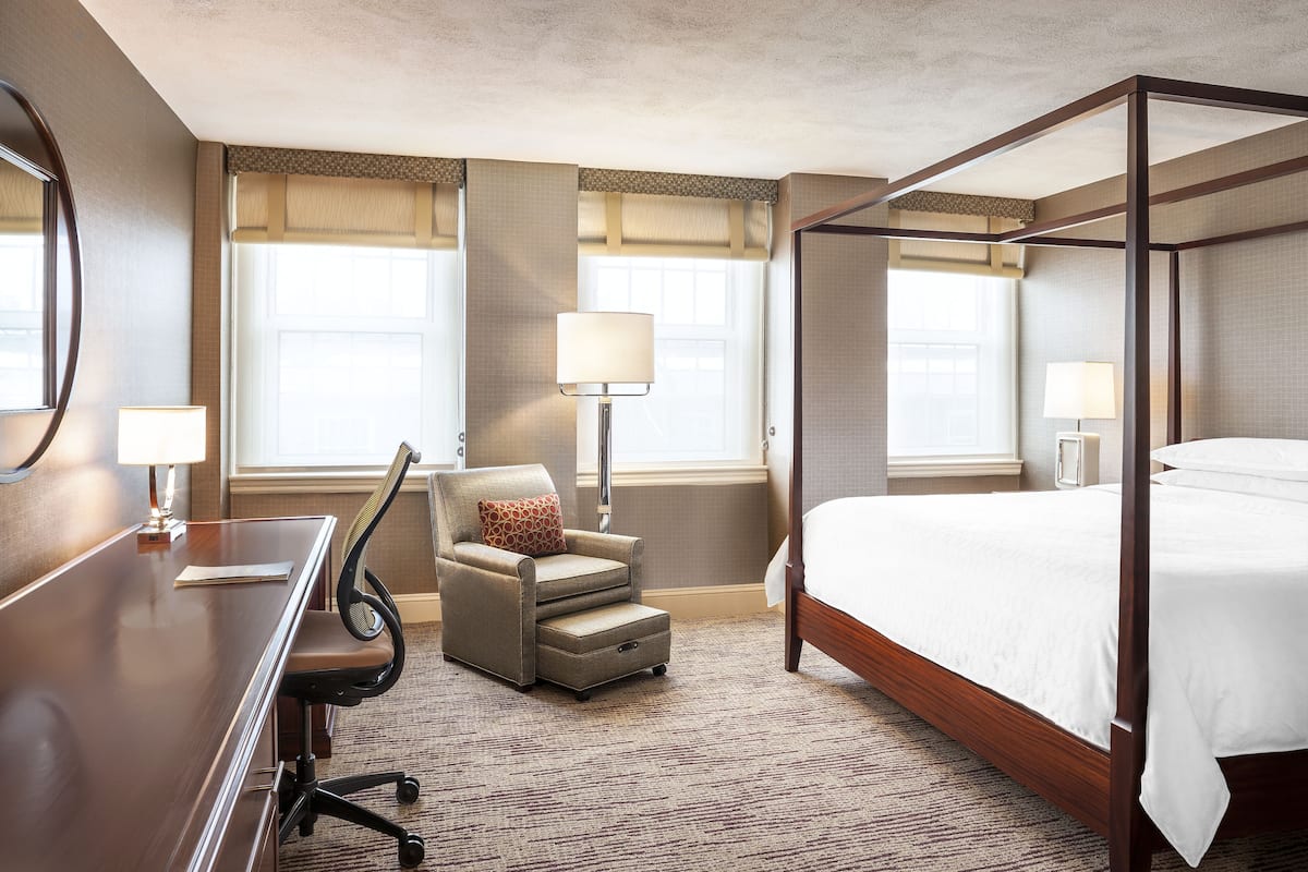 A hotel room with a four-poster bed, a desk with a chair, and two windows with curtains.
