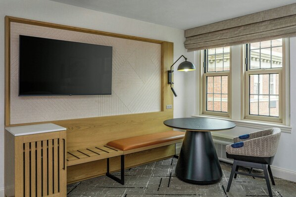 A modern room with a wooden bench, a round table, a TV mounted on the wall, and a window with a view of buildings.