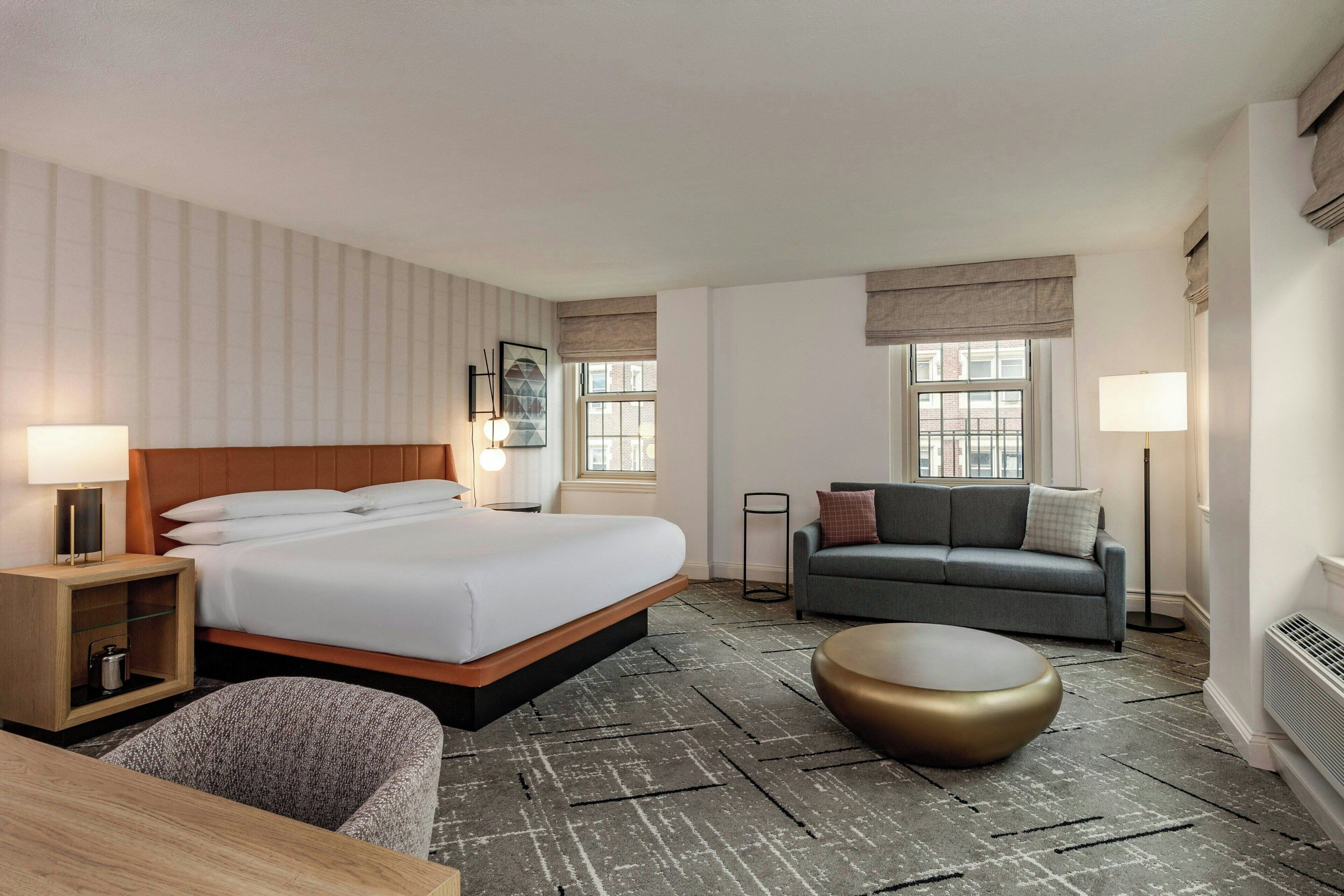 Une chambre d’hôtel comprenant un lit, un canapé, un pouf rond, une table de chevet et une lampe.