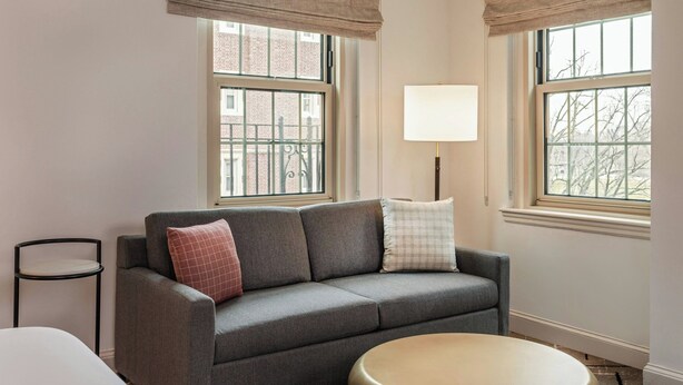 A room with a grey sofa, a round gold table, and two windows with a view of buildings. - Sheraton Commander Hotel (Cambridge)