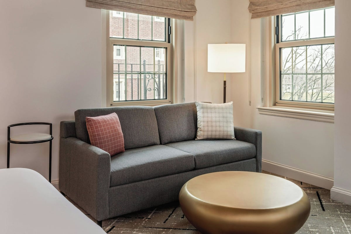 A room with a grey sofa, a round gold table, and two windows with a view of buildings.