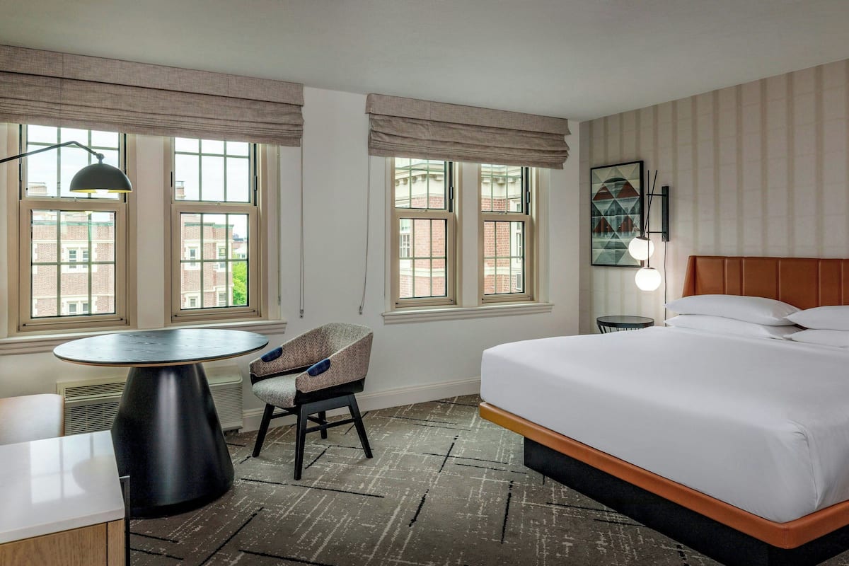 A hotel room with a large bed, a round table, a chair, and a view of buildings through the windows.
