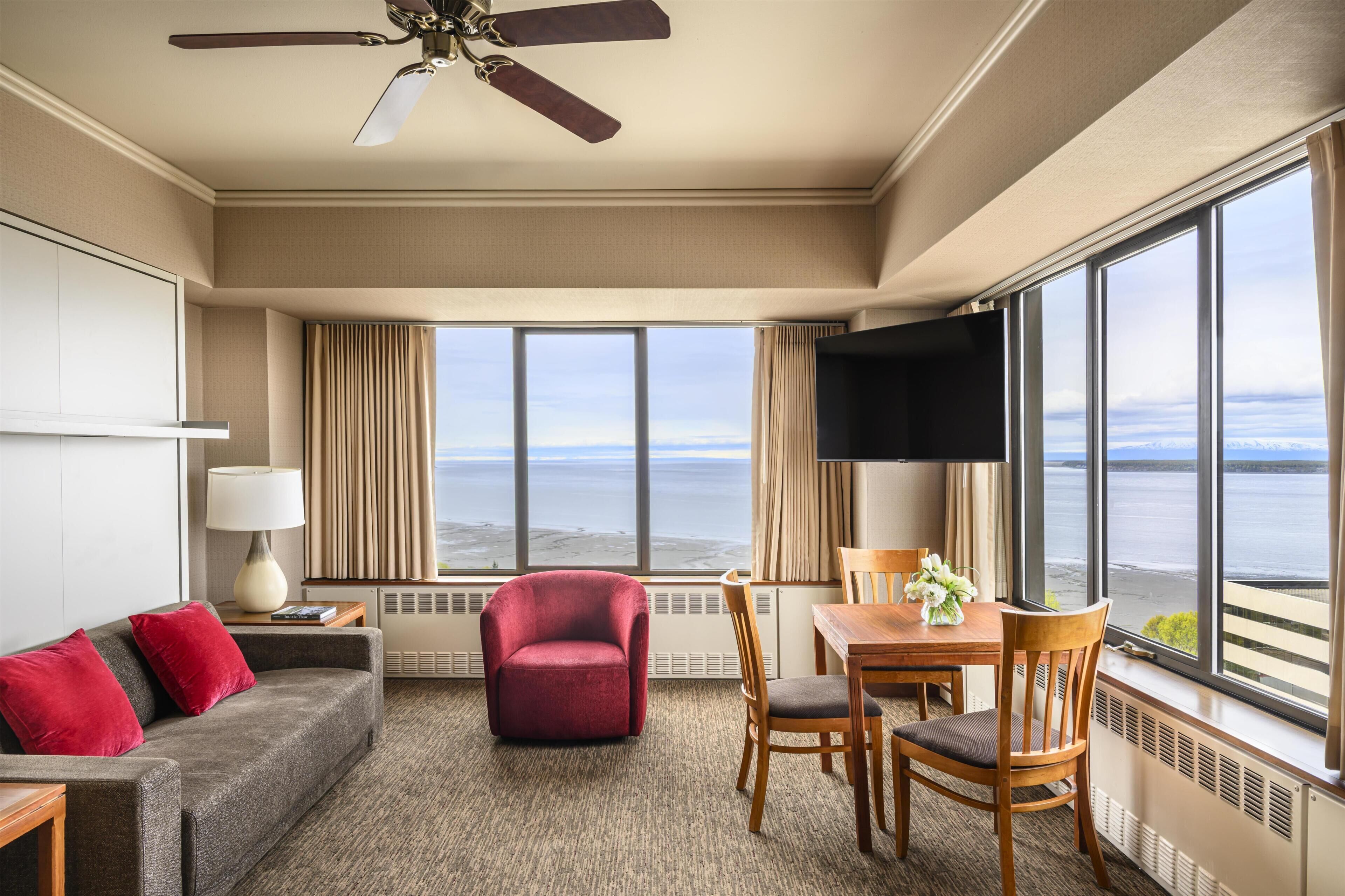 A hotel room with a large window offering a view of the sea, a ceiling fan, a red armchair, a wooden dining table with chairs, and a grey sofa with red cushions.