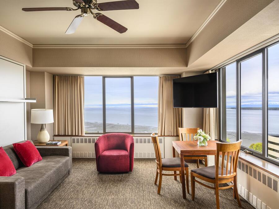 A hotel room with a large window offering a view of the sea, a ceiling fan, a red armchair, a wooden dining table with chairs, and a grey sofa with red cushions.