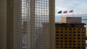 A view through a window with a mesh screen, featuring a building with multiple flags, including the UK and US flags, and a body of water in the background. - Hotel Captain Cook (Anchorage)