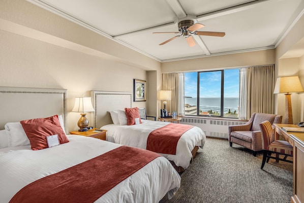 A hotel room with two beds, a desk, a chair, a ceiling fan, and a large window with a view of the ocean.