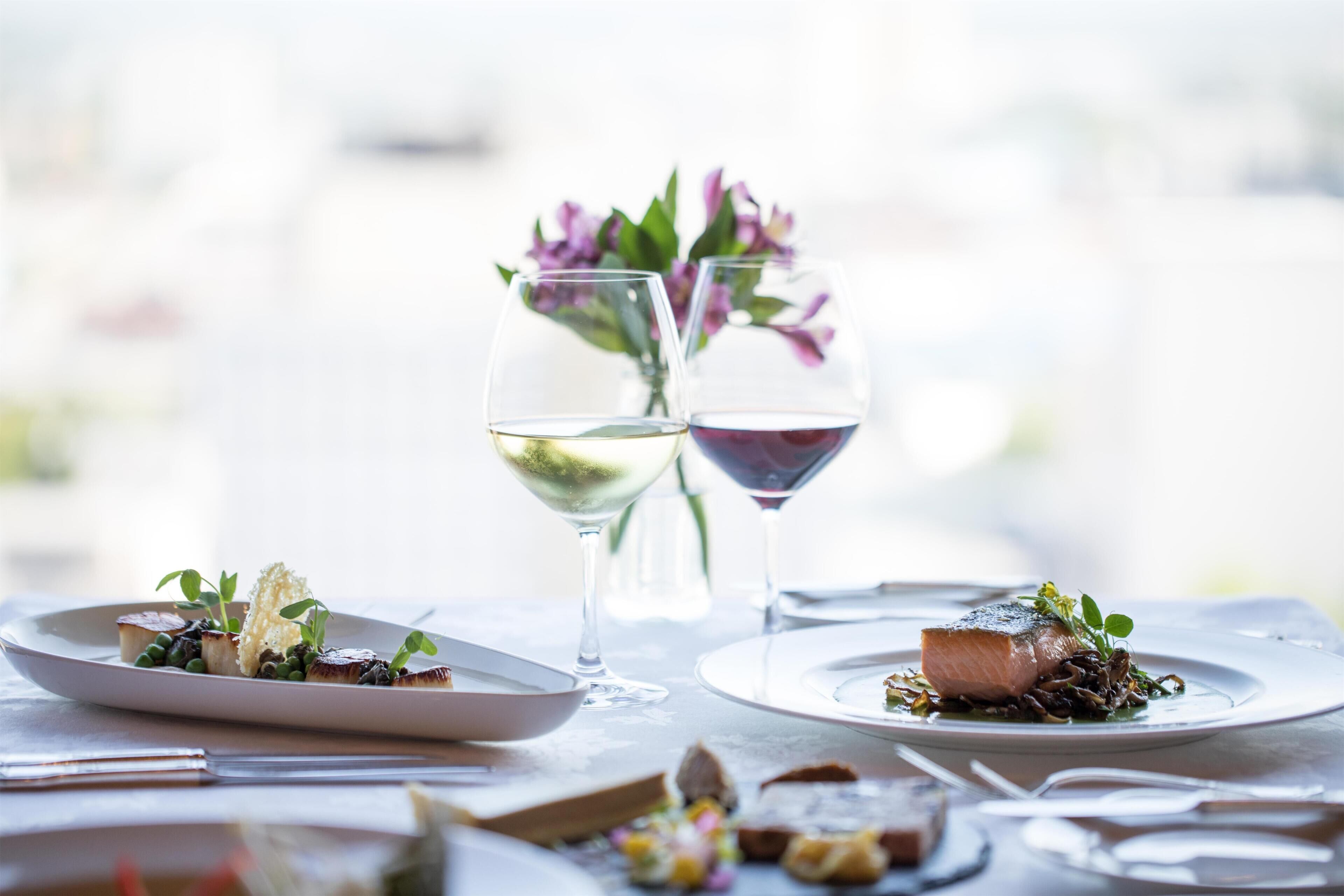 A dining table with plates of food, two wine glasses, and a vase with flowers.