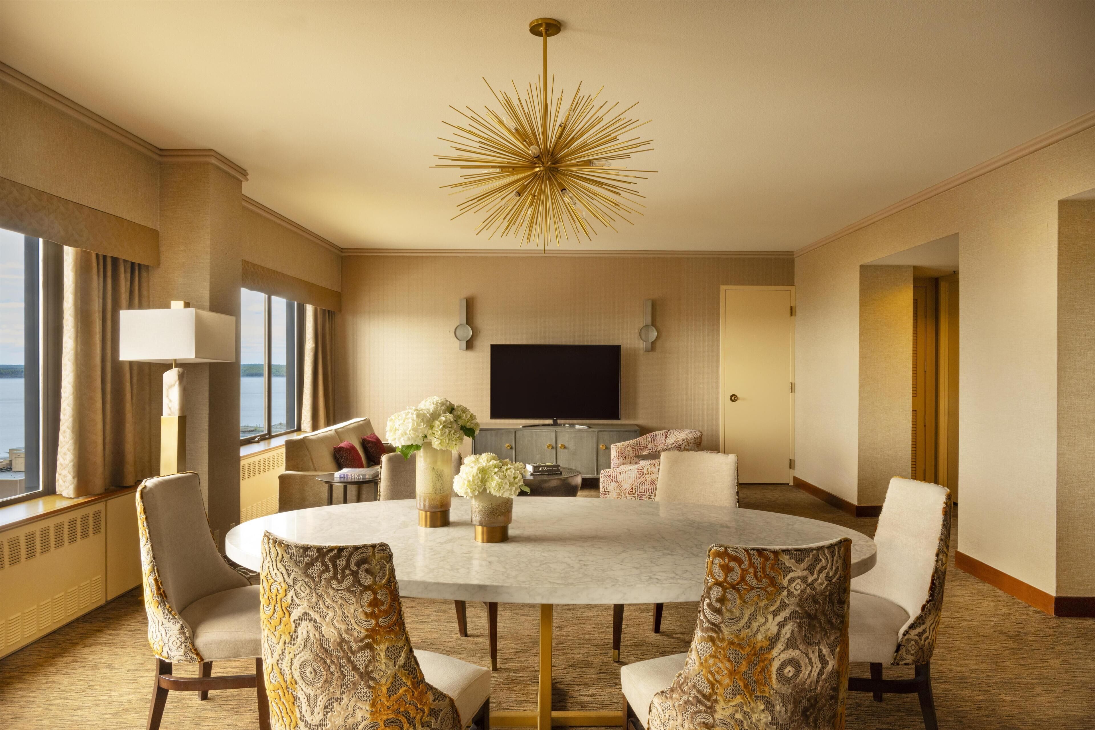 A hotel room with a round dining table, patterned chairs, a television, and a large window.