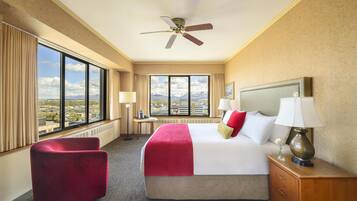 A hotel room with a bed, a red chair, a nightstand, a lamp, and a view of the city and mountains.