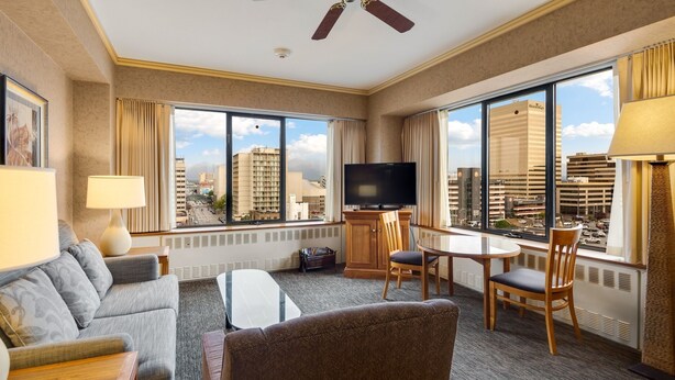 A hotel room with a large window, a ceiling fan, a flat-screen TV, a sofa, a coffee table, and a dining table with chairs. - Hotel Captain Cook (Anchorage)