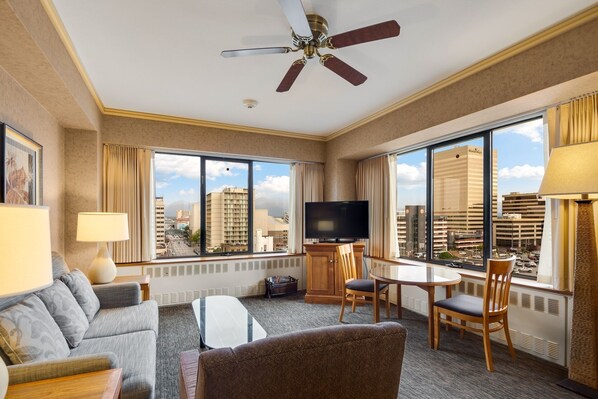 Una habitación de hotel con un ventanal grande, ventilador de techo, televisor de pantalla plana, sofá, mesa de centro y una mesa de comedor con sillas.