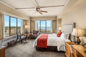A hotel room with a bed, a desk, a chair, a table, a ceiling fan, and large windows with a view of the city and sea. - Hotel Captain Cook (Anchorage)