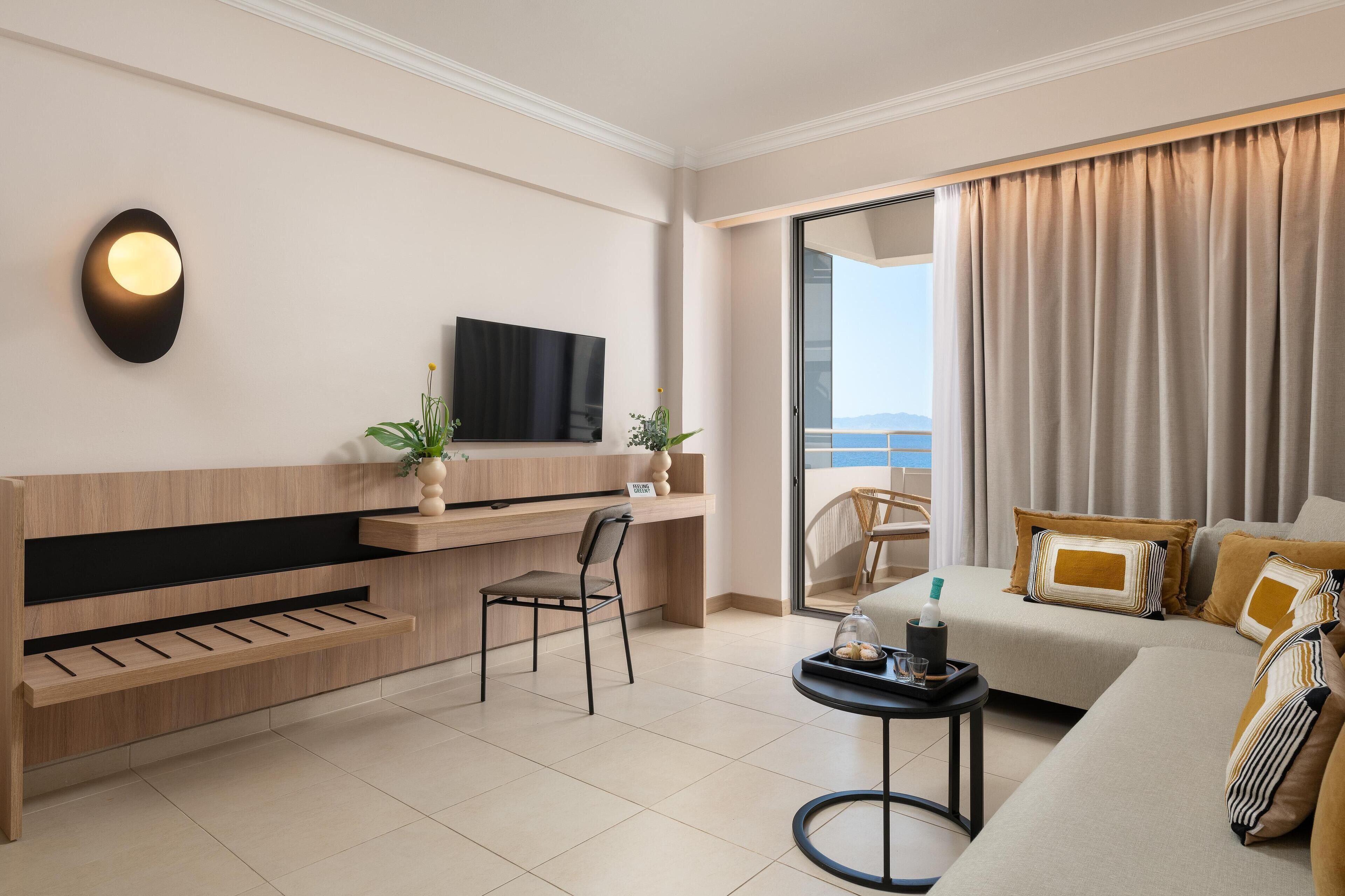 Ein modernes Hotelzimmer mit einem Wand-Fernseher, einem Holzkonsolentisch mit Stuhl, einer Couch mit Zierkissen und einem Balkon mit Aussicht.