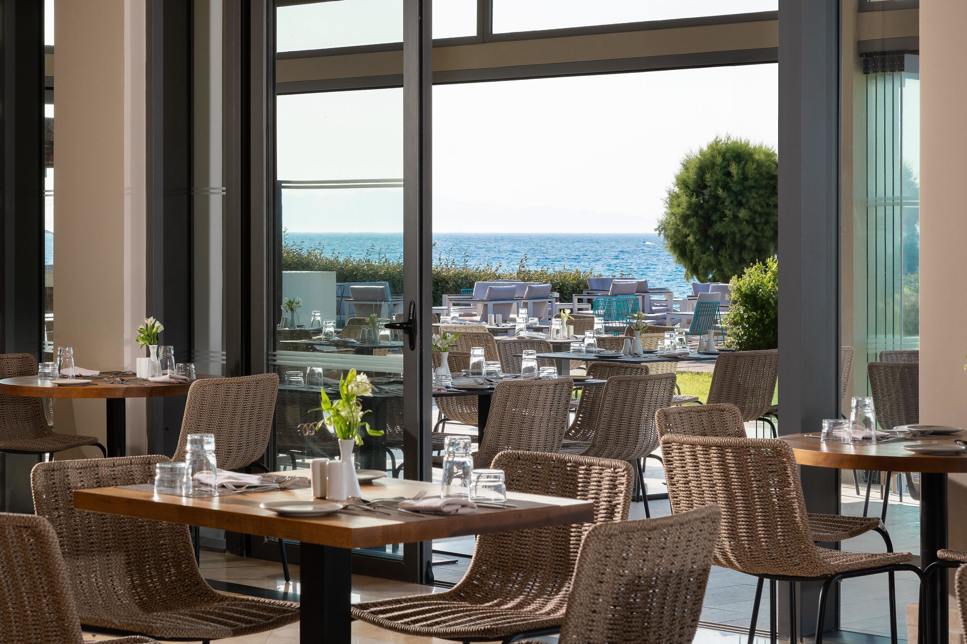 En restaurant med fletstole og -borde, store vinduer med havudsigt og udendørs siddepladser.