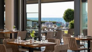 En restaurant med fletstole og -borde, store vinduer med havudsigt og udendørs siddepladser.
