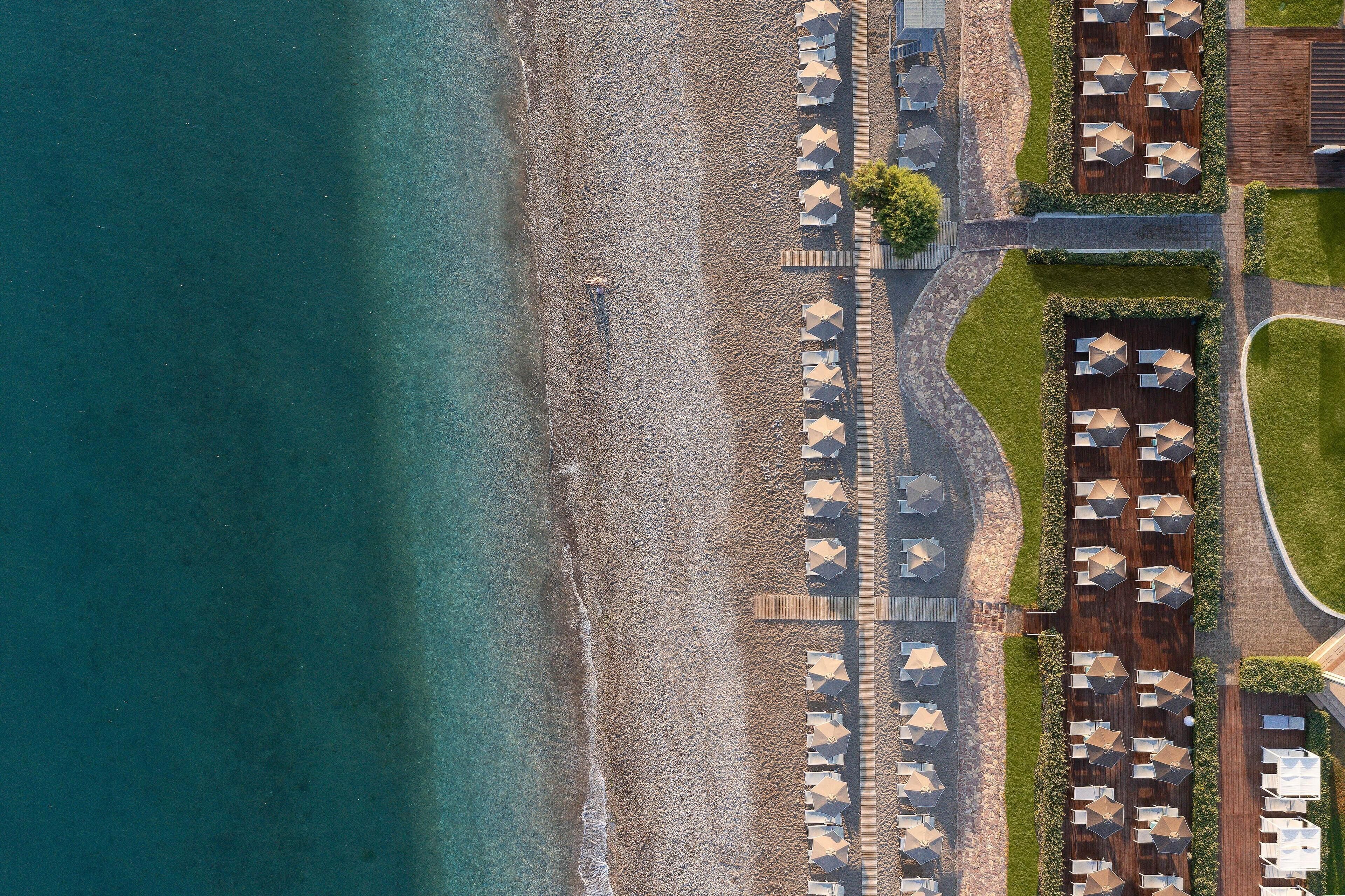 Ein Luftbild eines Strandanliegers mit Reihen von Sonnenschirmen und Liegestühlen, einer Holzpromenade und einem gepflasterten Weg entlang des Strandes.